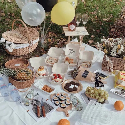 An overhead shot of a diverse picnic spread on a white blanket, including sushi rolls, pineapple, various berries, and champagne. The assortment of food and drinks captures a modern, curated French picnic style.