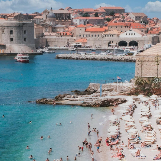 Crowded beach beneath Dubrovnik's medieval city walls with crystal-clear Adriatic waters, exemplifying Croatia's unique culture and beach combination