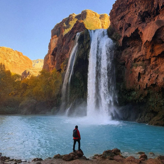 A person stands on a rock admiring the vibrant turquoise water of Havasu Falls, surrounded by red rock canyons.