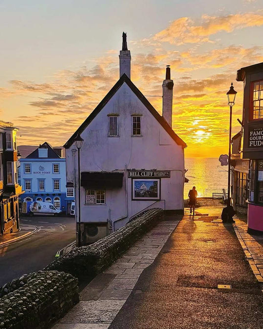 A picturesque sunset scene in Lyme Regis, with a white building and street leading towards the golden sea and sky, silhouetting figures and streetlights.