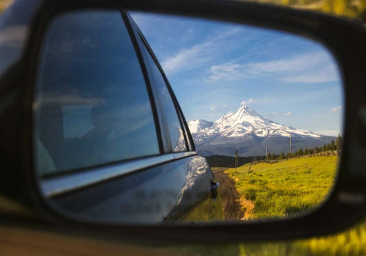 Snow-capped mountain peak reflected in car side mirror during road trip to hiking destination, representing the journey to discover the best hiking snacks for alpine adventures