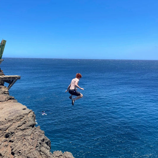 Cliff jumping action showing person jumping feet first from rocky cliff into clear blue ocean water with another swimmer below demonstrating cliff jumping technique for beginners