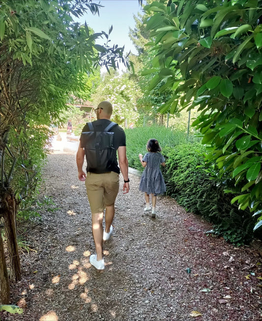 Modern father wearing stylish waterproof backpack walking hand-in-hand with young daughter on garden path surrounded by lush greenery