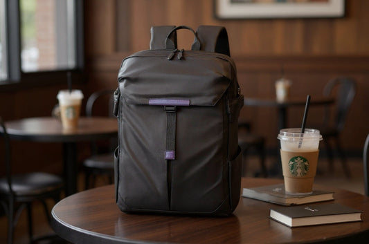 Black Backpackbeat 20L commuter backpack placed on a coffee shop table next to iced coffee and books, showing a calm, everyday work routine.