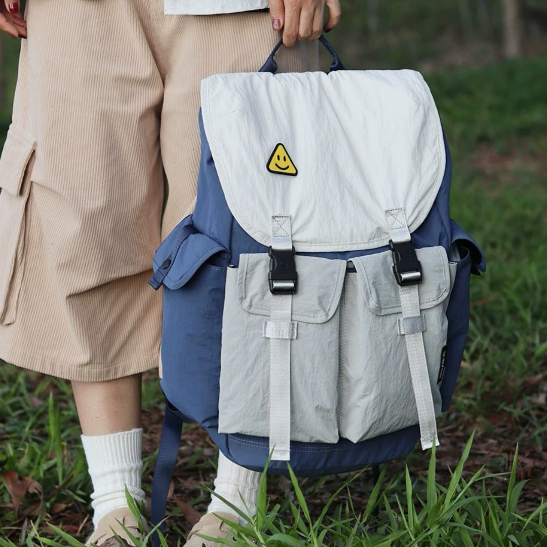 Hand holding blue-gray lightweight waterproof backpack displaying front organization pockets and signature smiley patch