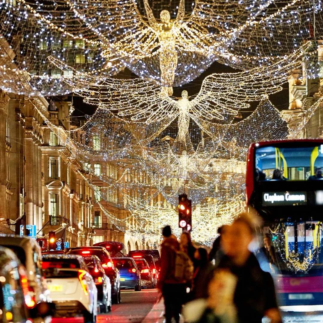 Large illuminated angel sculptures made of white lights suspended over busy city street with traffic and pedestrians taking photos during Christmas season