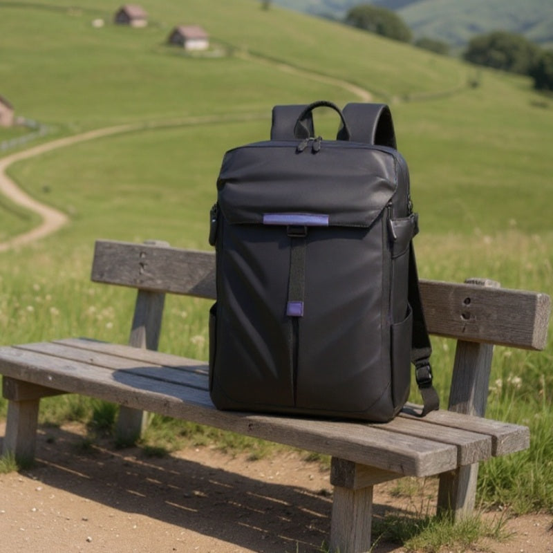 A black waterproof backpack resting upright on a wooden bench in a grassy park, with rolling hills and small houses in the background.