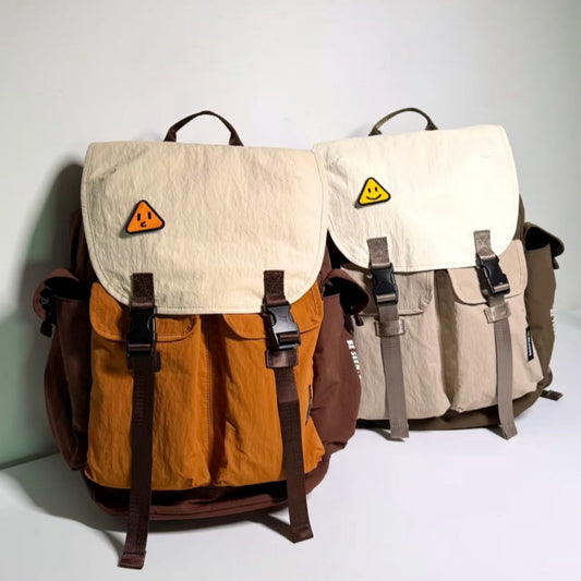 Two Backpackbeat 7706 waterproof backpacks in orange-brown and beige-green color combinations, both featuring triangle smiley patches and earth tone designs