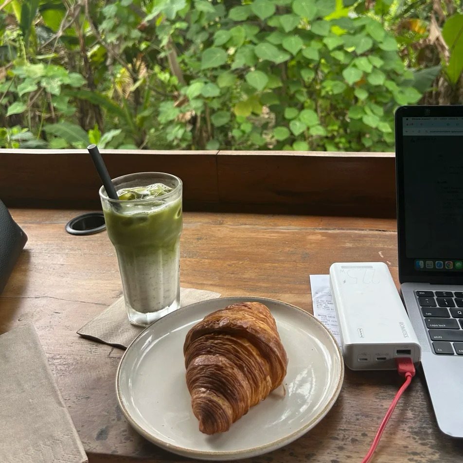 Croissant and iced matcha latte on wooden table with laptop and power bank - tropical coworking cafe breakfast scene