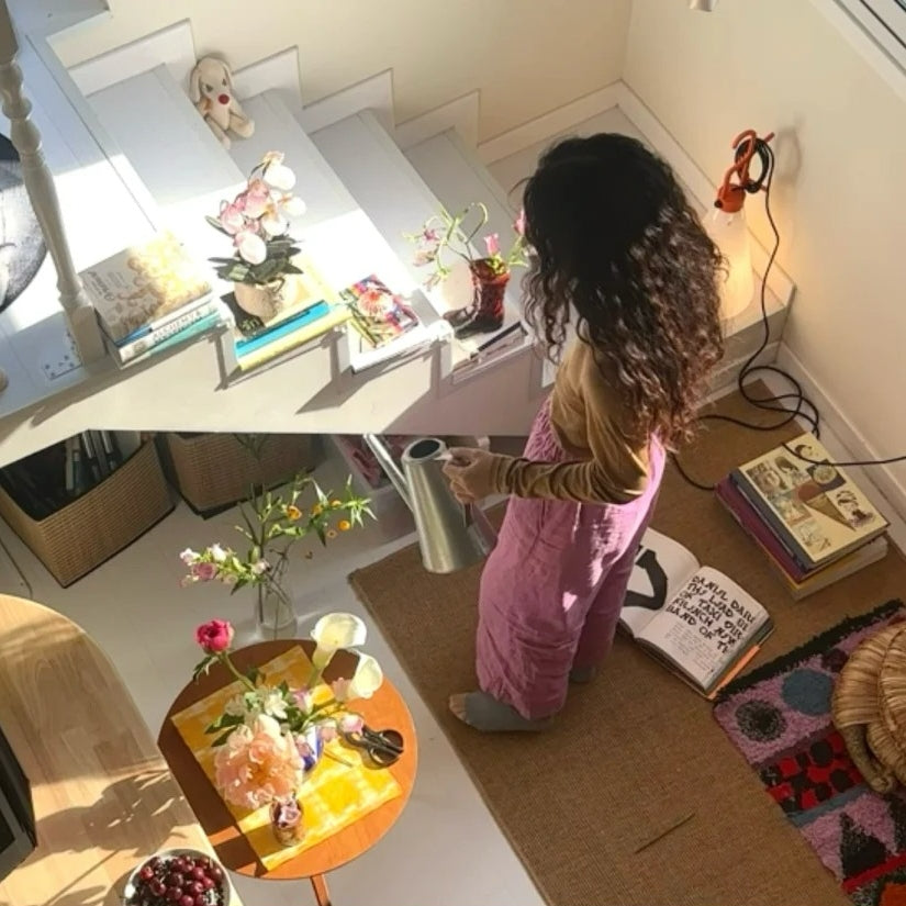 Woman watering plants in sunny apartment workspace - digital nomad lifestyle home office with books, flowers, and natural light