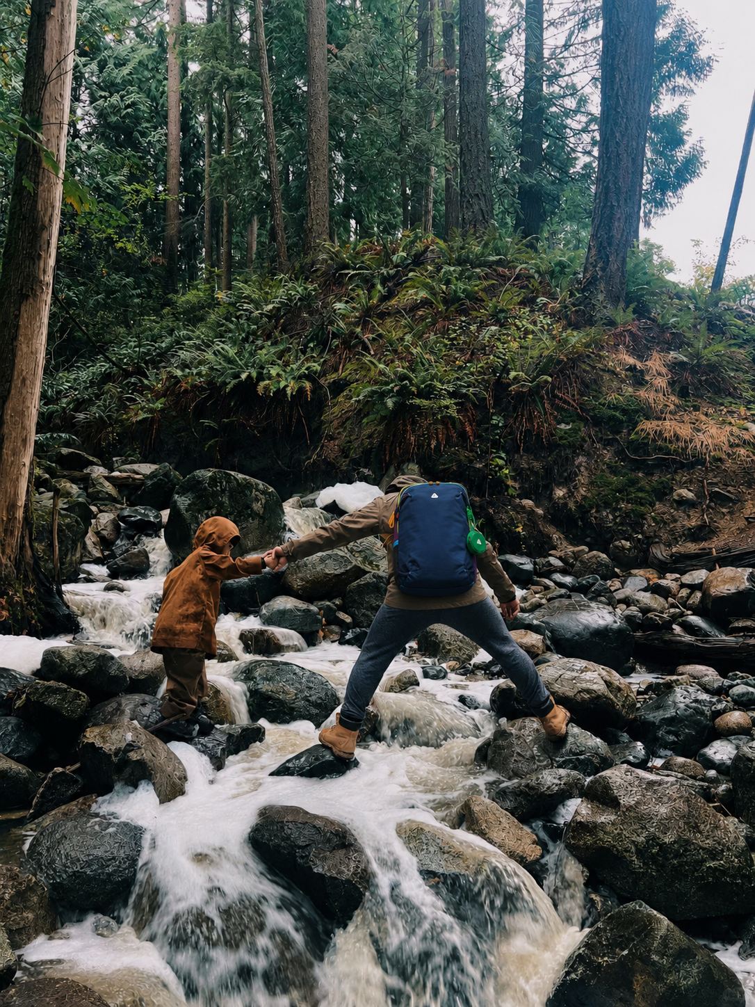 Parent carrying a blue hiking backpack while helping a child cross a flowing rocky stream in the forest.