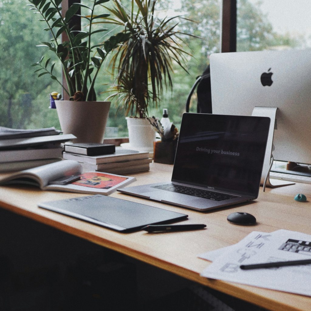A tidy wooden desk with an open MacBook, drawing tablet, stacked books and two potted plants in front of large windows — a typical work-from-anywhere setup