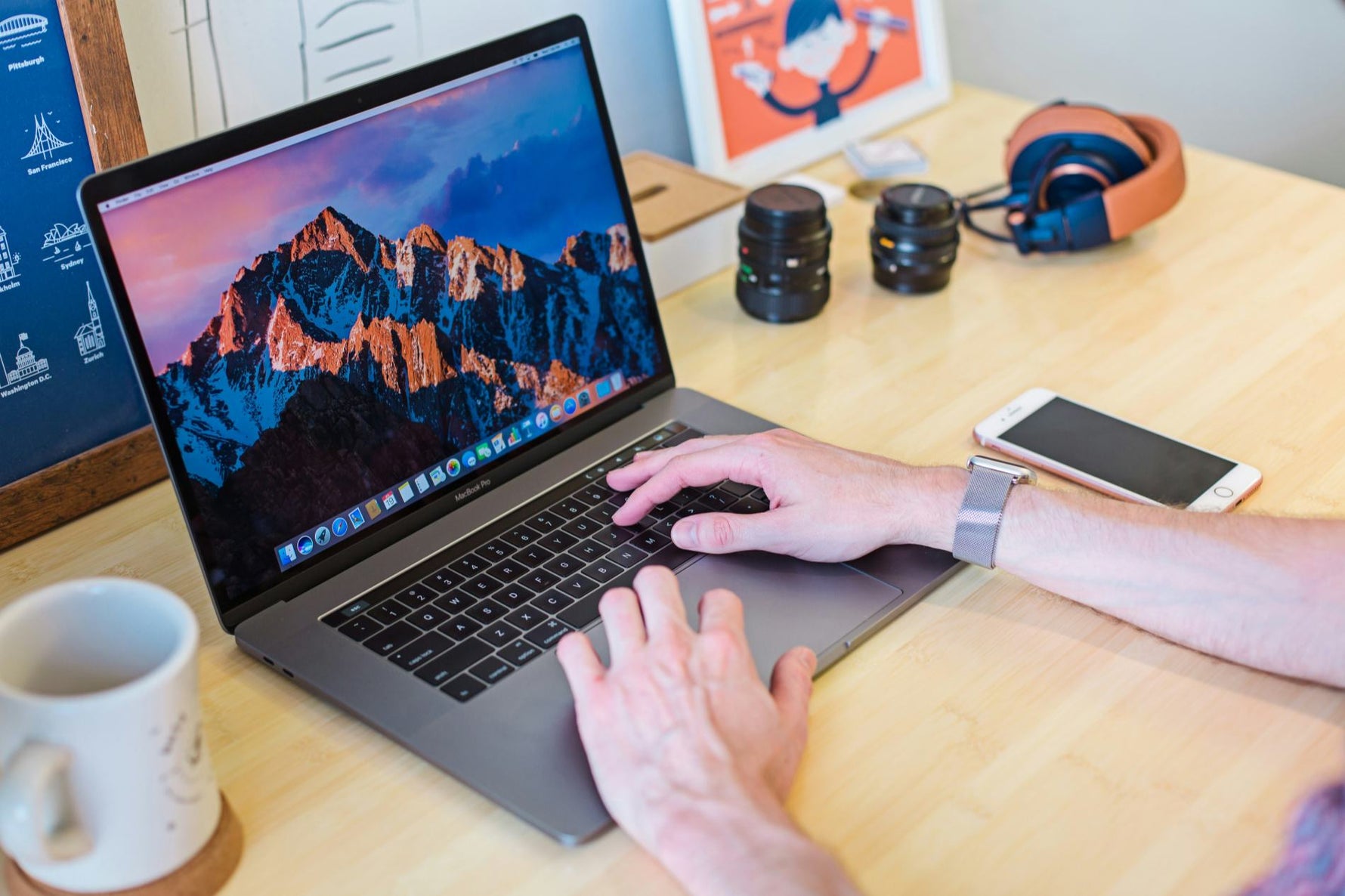 Hands typing on a MacBook Pro at a wooden desk with camera lenses, headphones, a phone and a coffee mug — a remote work setup ready to go anywhere