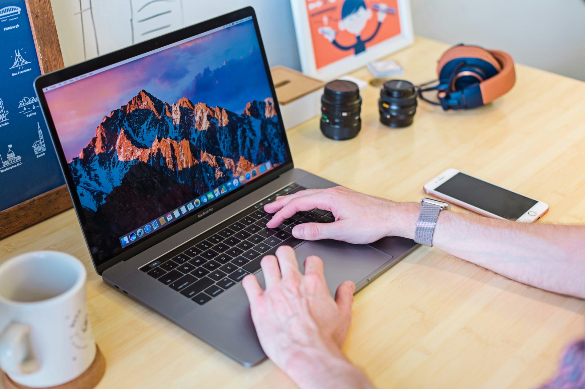 Hands typing on a MacBook Pro at a wooden desk with camera lenses, headphones, a phone and a coffee mug — a remote work setup ready to go anywhere