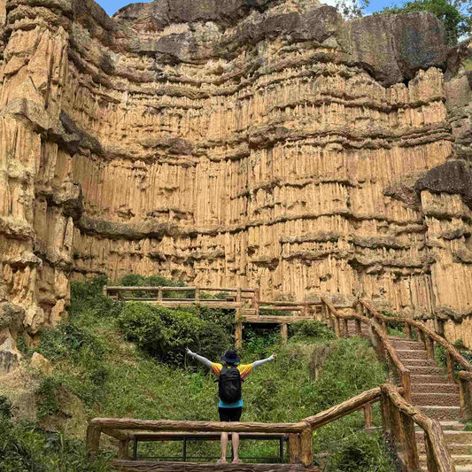 Solo female traveler wearing yellow jacket and Backpackbeat 7705 waterproof backpack standing at ancient canyon cliff with arms spread wide, demonstrating best travel accessories for outdoor adventures and hiking