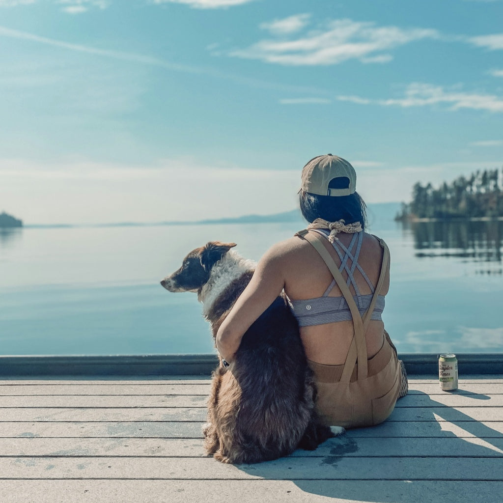 Back view of a young woman wearing a cap and overalls sitting on a wooden dock, hugging her fluffy border collie dog while looking out at a calm, wide lake under a blue sky.