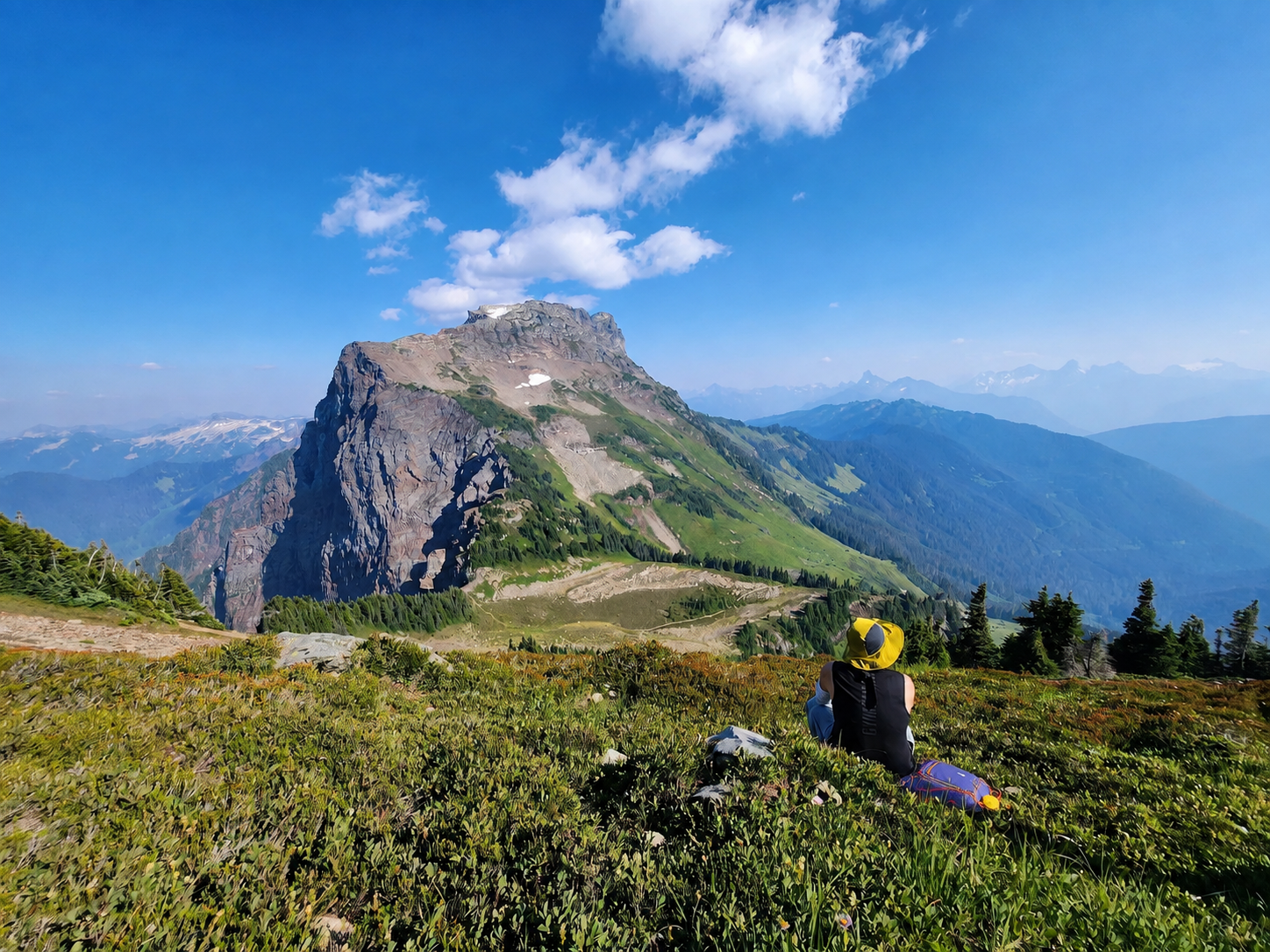 Hiker sitting in an alpine meadow and looking toward a rocky mountain beneath a blue sky.
