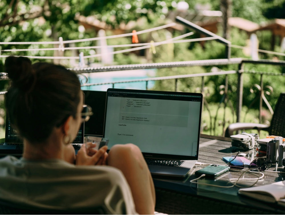 Woman working on code from an outdoor table surrounded by gear like a drone, sunglasses, and phone chargers, freelancing before committing to full-time nomad life
