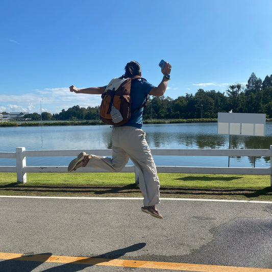 Person wearing Backpackbeat 7705 multicolor canvas backpack jumping joyfully near waterfront, demonstrating lightweight travel backpack for outdoor adventures
