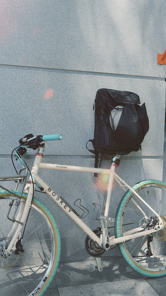 Expandable black backpack hanging on a white bicycle against a grey city wall, helmet tucked into the front pocket, ready for everyday urban riding.