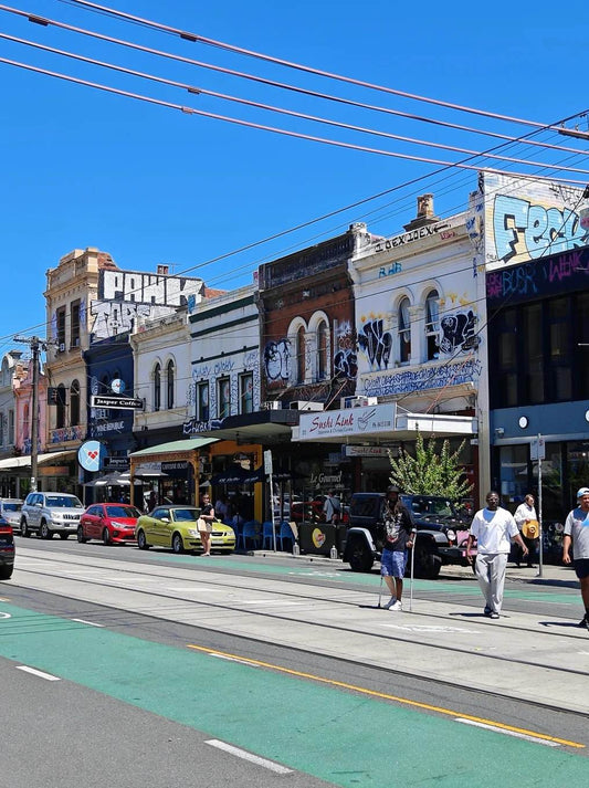 Colorful street view of Brunswick St. in Fitzroy, lined with shops, graffiti, and a tram line under bright blue sky