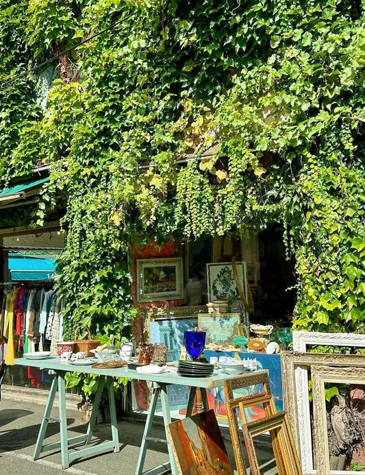 A vine-covered shopfront at Saint-Ouen flea market, displaying vintage frames and oil paintings on tables