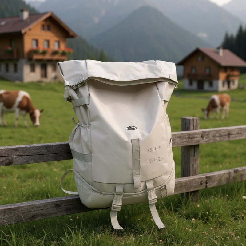 Beige 8803 waterproof backpack 34L positioned on wooden fence in Alpine countryside with traditional chalets and grazing cattle, showcasing outdoor adventure durability