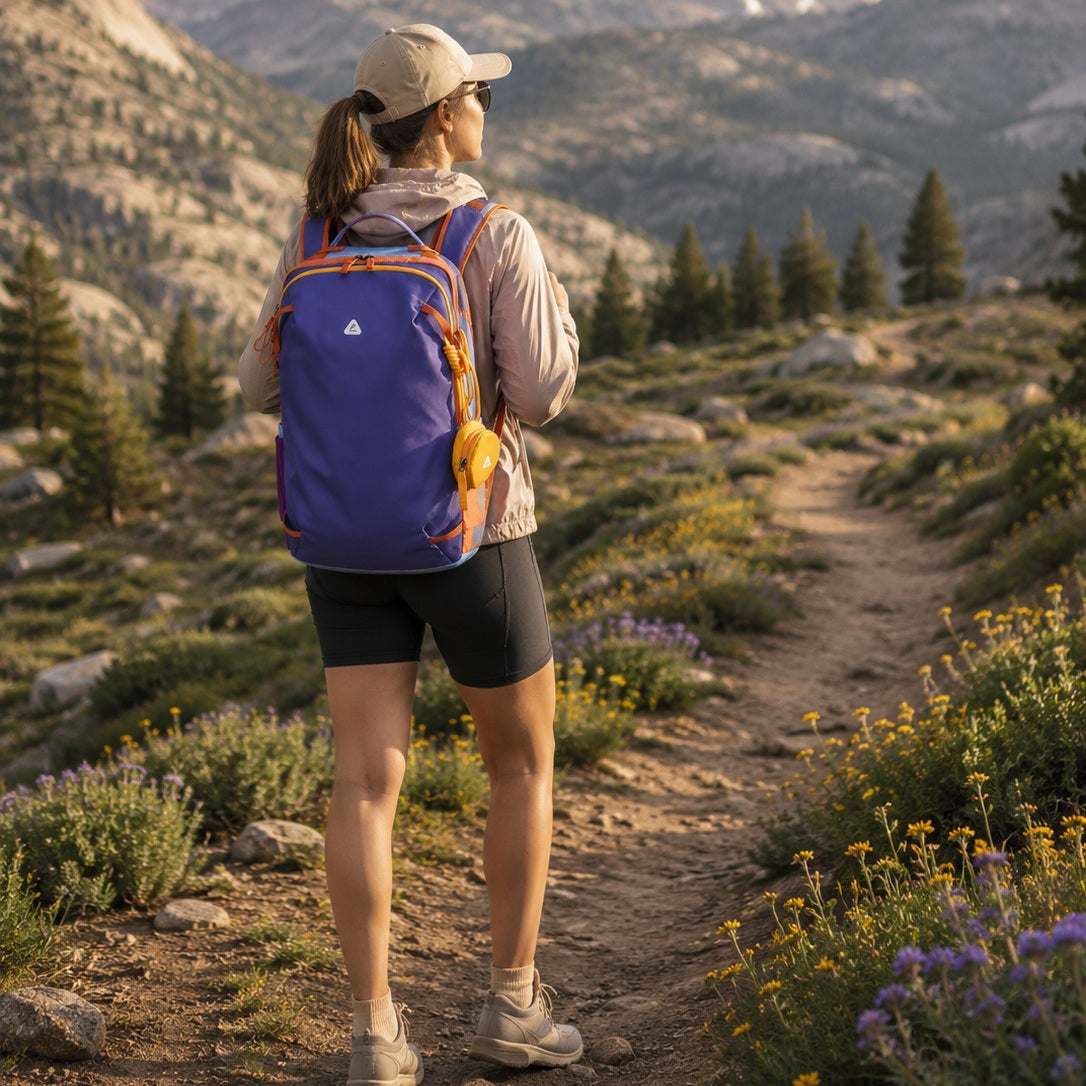 Woman hiking on a scenic mountain trail wearing the BackpackBeat 8809 Lightweight Hiking Backpack 19L in purple with orange trim and yellow accessory pouch during a summer outdoor adventure.