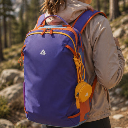 Close-up rear view of the BackpackBeat 8809 Lightweight Hiking Backpack 19L featuring a purple body, orange trim, yellow zipper accents and a detachable yellow accessory pouch on a forest hiking trail.