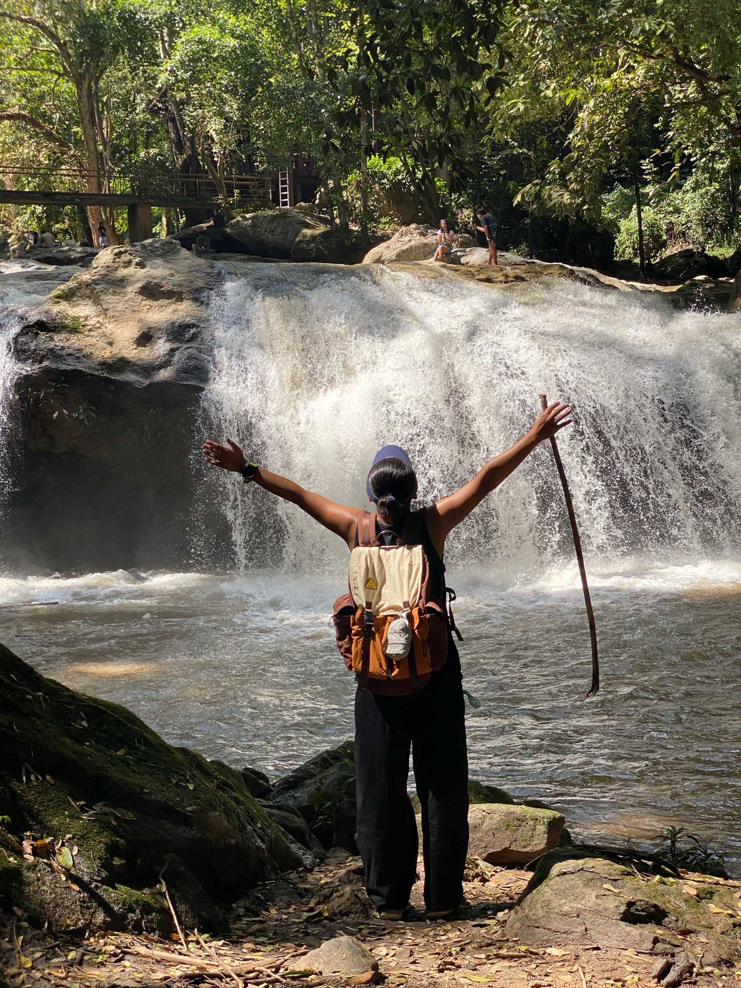A woman stands with arms outstretched, holding a hiking stick, facing a large, cascading waterfall. She is wearing the Voyager 7706 Lightweight Stylish Waterproof Backpack 26L, highlighting its suitability as one of the Best Lightweight Travel Backpacks for Women 2026.