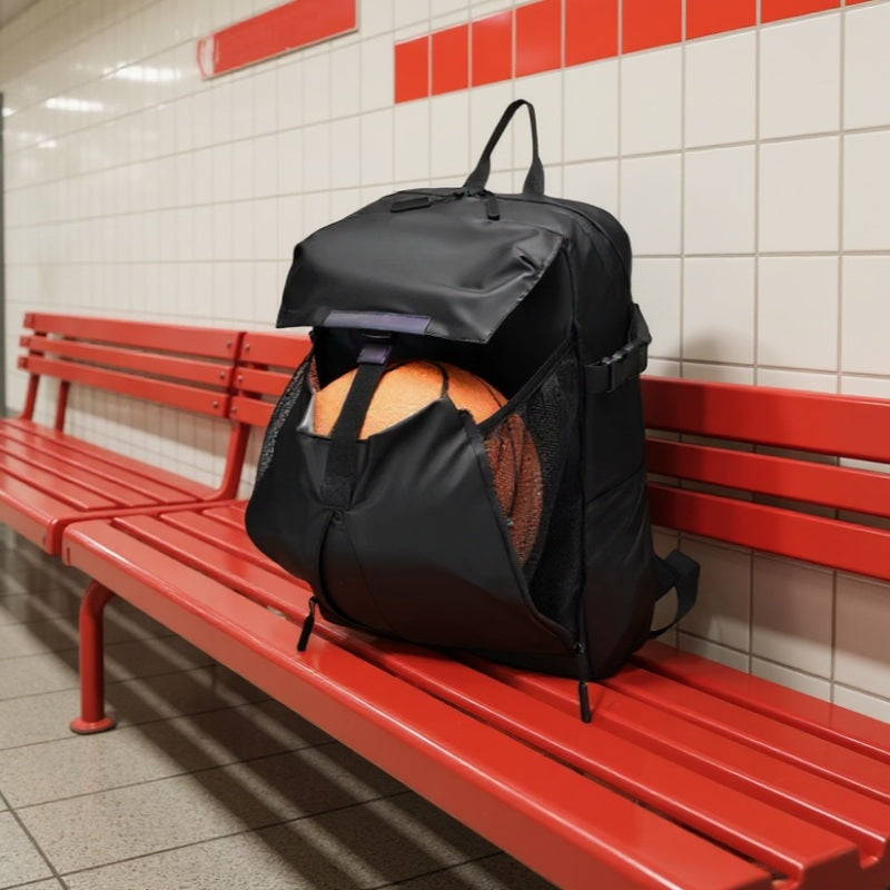 A black waterproof backpack with a basketball in its mesh pocket placed on a red bench inside a tiled subway station with red and white walls.